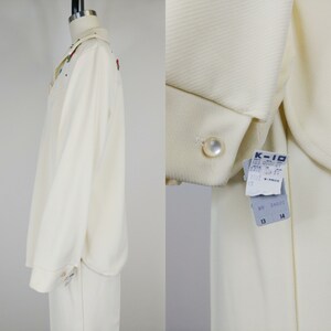 Peut inclure: Un ensemble chemise et pantalon blanc &agrave; manches longues avec un bouton en perle et des fleurs brod&eacute;es. La chemise a un col et le pantalon a une jambe droite. L'&eacute;tiquette indique "K-10 103 000 37 372 13 17 1002" et "RN 34030 13 14".