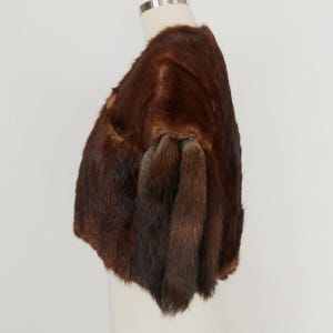 May include: A brown fur bolero jacket displayed on a mannequin. The jacket has a rounded shape and features a rich, dark brown color with varying shades. The fur appears soft and luxurious, with a natural texture. The bolero has short sleeves.