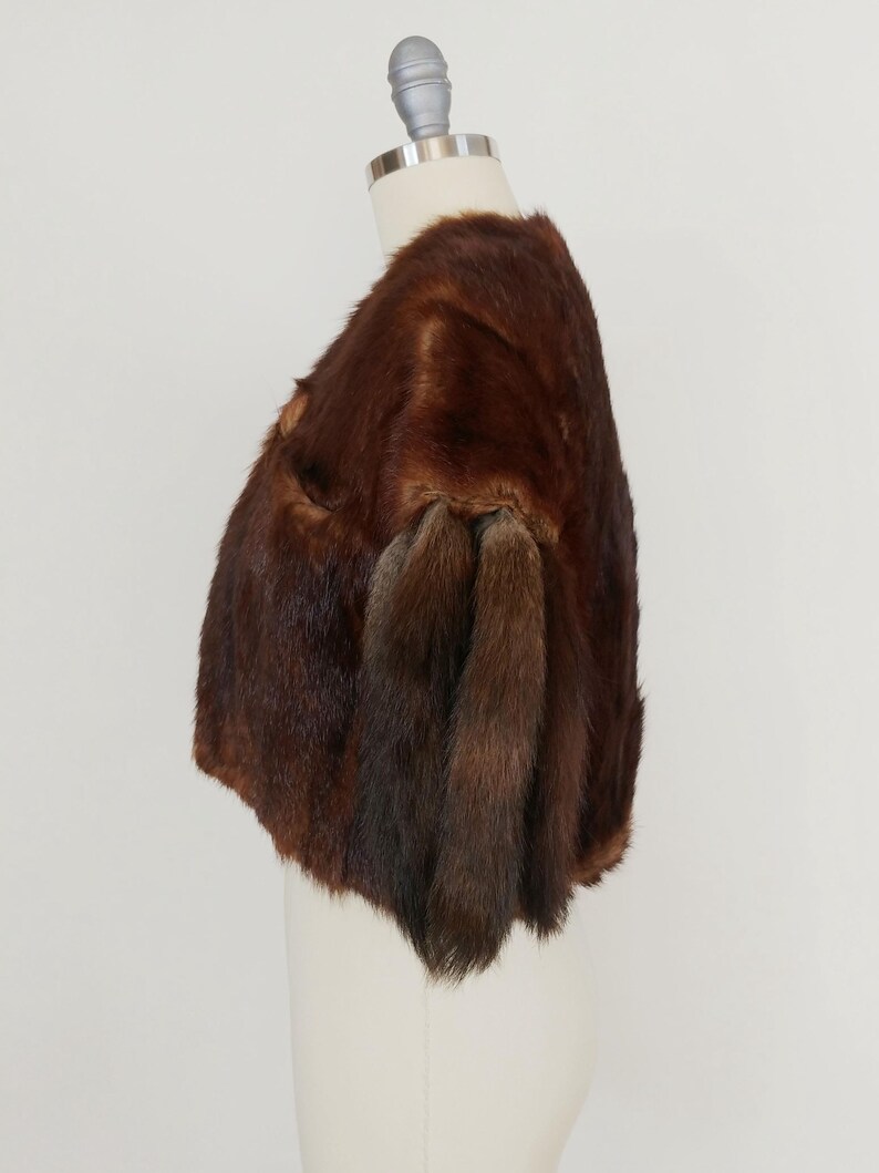 May include: A brown fur bolero jacket displayed on a mannequin. The jacket has a rounded shape and features a rich, dark brown color with varying shades. The fur appears soft and luxurious, with a natural texture. The bolero has short sleeves.