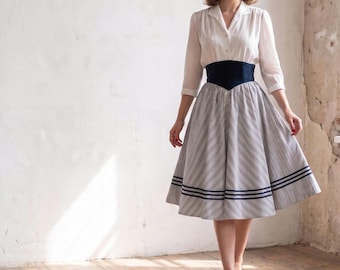 May include: A two-piece outfit consisting of a long-sleeved white button-down blouse with a collar and a high-waisted, full-length skirt featuring a diagonal pinstripe pattern, a wide dark blue waistband, and two horizontal dark blue stripes at the hem.