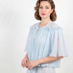 Könnte beinhalten: Ein hellblaues, transparentes, plissiertes Capelet mit einem gerüschten Kragen und einem zarten Bindeband am Ausschnitt. Das Kleidungsstück hat weite, fließende Ärmel und eine weiche, halbtransparente Textur, die über Schultern und Oberkörper fällt.