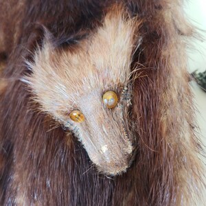 May include: Close-up of a brown fur item with a stylized animal face. The face is made of lighter brown fur with two amber-colored eyes and a snout. The surrounding fur is a darker shade of brown.