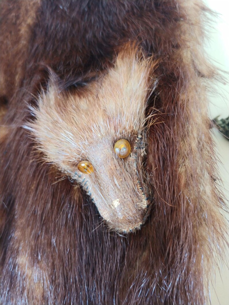 May include: Close-up of a brown fur item with a stylized animal face. The face is made of lighter brown fur with two amber-colored eyes and a snout. The surrounding fur is a darker shade of brown.