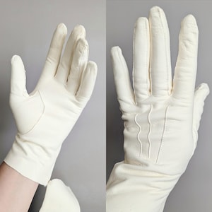 May include: Off-white leather gloves. The gloves are shown from two angles, one showing the front with a reinforced palm, and the other showing the back with decorative stitching. The gloves are a classic style.