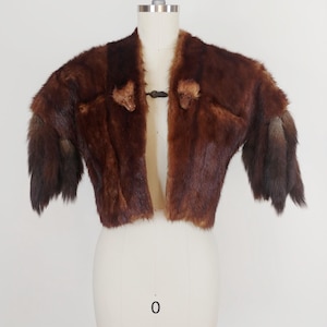 May include: A vintage brown fur bolero jacket displayed on a mannequin. The jacket features a dark brown color with a textured appearance and a hook closure. The sleeves have a darker shade of brown at the ends.