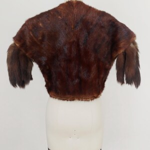 May include: A vintage brown fur bolero jacket displayed on a mannequin. The jacket features a rounded neckline and short sleeves. The fur appears to be a rich, dark brown color with variations in tone, giving it depth and texture. The garment is cropped at the waist.