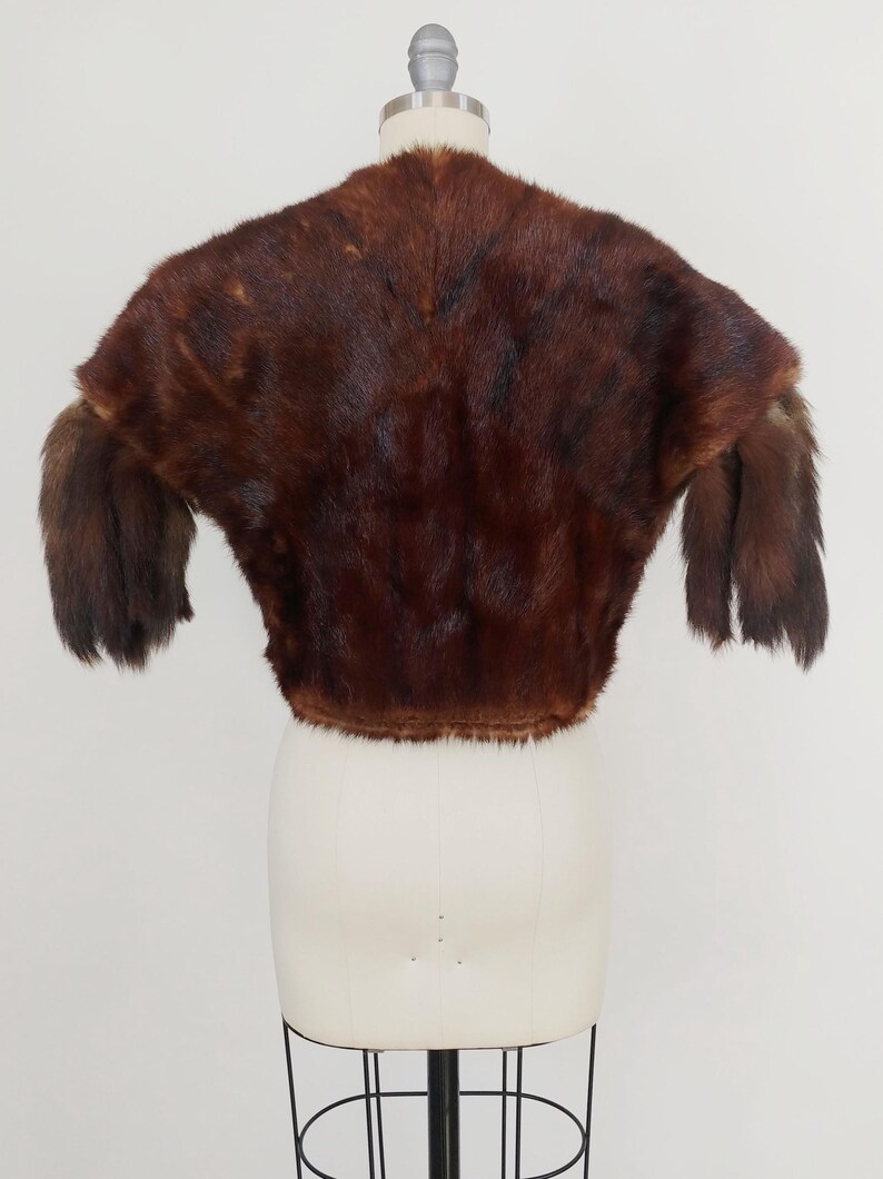 May include: A vintage brown fur bolero jacket displayed on a mannequin. The jacket features a rounded neckline and short sleeves. The fur appears to be a rich, dark brown color with variations in tone, giving it depth and texture. The garment is cropped at the waist.