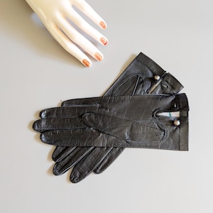 May include: A pair of black leather gloves featuring a smooth texture, stitched detailing on the fingers, and a small metallic button closure at the wrist. The gloves are positioned overlapping each other, showcasing the wrist opening and seams.
