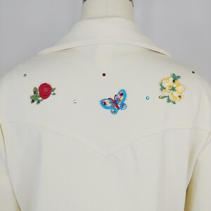 Peut inclure: Un blazer blanc avec des fleurs brod&eacute;es et un papillon au dos. Les fleurs sont rouges, jaunes et vertes. Le papillon est bleu et rouge.