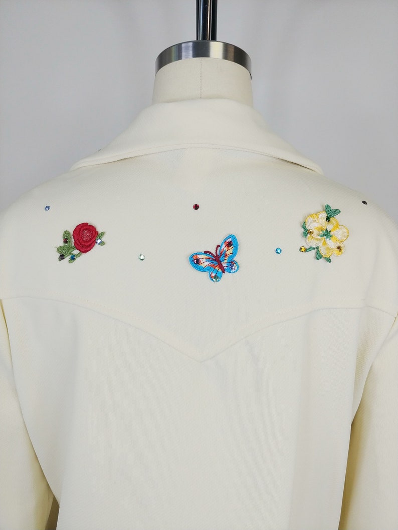 Peut inclure: Un blazer blanc avec des fleurs brod&eacute;es et un papillon au dos. Les fleurs sont rouges, jaunes et vertes. Le papillon est bleu et rouge.
