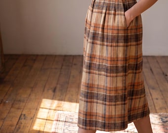 May include: A midi-length skirt featuring a plaid pattern in shades of tan, brown, and dark blue. The skirt has a high-waisted design with a waistband, belt loops, and vertical pleats at the waistline. It includes side pockets and a straight hem.