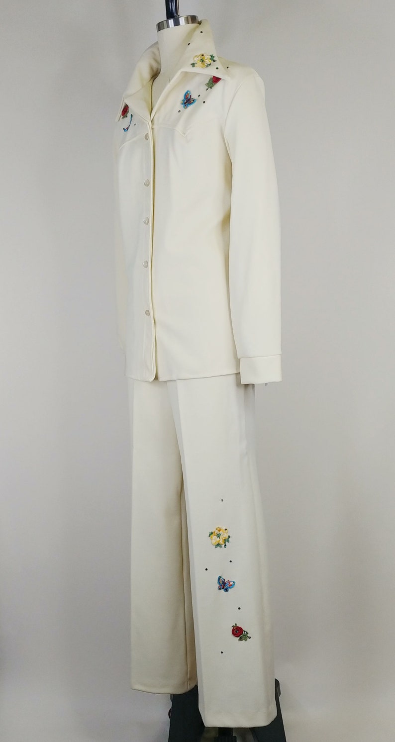 Peut inclure: Un tailleur pantalon blanc avec des papillons et des fleurs brod&eacute;s. La veste a un devant boutonn&eacute; et des manches longues. Le pantalon a une jambe large.