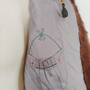 May include: Close-up of the inside of a vintage fur coat. The coat's lining is a light gray with vertical stripes and a pocket embroidered with a green bow and pink flowers. The fur is a reddish-brown color.