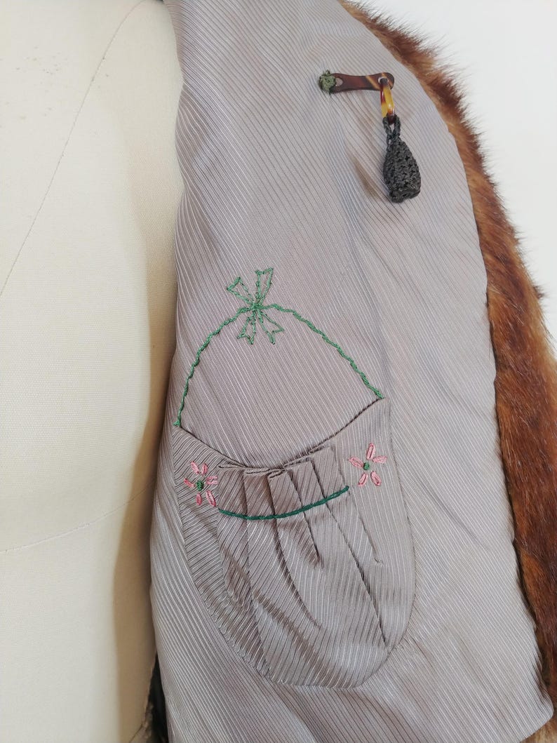 May include: Close-up of the inside of a vintage fur coat. The coat's lining is a light gray with vertical stripes and a pocket embroidered with a green bow and pink flowers. The fur is a reddish-brown color.