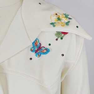 Peut inclure: Une chemise blanche &agrave; col avec des fleurs brod&eacute;es et un papillon sur le col. Les fleurs sont jaunes, rouges et vertes, et le papillon est bleu, rouge et jaune. Il y a aussi de petits strass &eacute;parpill&eacute;s autour du col.