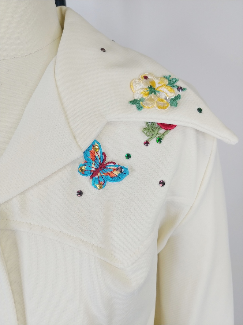 Peut inclure: Une chemise blanche &agrave; col avec des fleurs brod&eacute;es et un papillon sur le col. Les fleurs sont jaunes, rouges et vertes, et le papillon est bleu, rouge et jaune. Il y a aussi de petits strass &eacute;parpill&eacute;s autour du col.