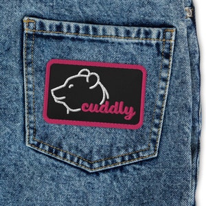 Puede incluir: Un parche bordado rosa y negro con un contorno blanco de la cabeza de un oso y la palabra "cuddly" en letras rosas. El parche está cosido en un bolsillo de mezclilla azul.