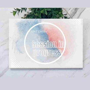 以下が含まれることがあります： 青とピンクの水彩画の背景を持つ白い看板。看板には、白い円の中に「静かに」と「セッション進行中」という白い文字が書かれています。