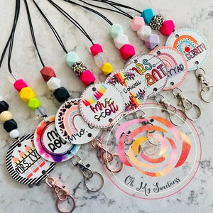 May include: Assortment of colorful teacher ID badge lanyards with rainbow and patterned designs. Each lanyard features a round badge with a teacher's name and a rainbow design. The lanyards are made with silicone beads and black string.