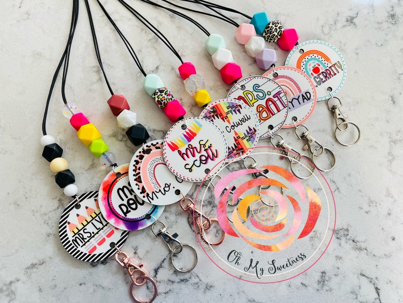 May include: Assortment of colorful teacher ID badge lanyards with rainbow and patterned designs. Each lanyard features a round badge with a teacher's name and a rainbow design. The lanyards are made with silicone beads and black string.