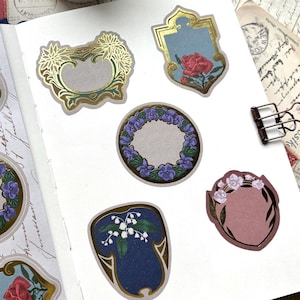 May include: A roll of adhesive paper labels and several individual stickers applied to a notebook page. The stickers feature ornate gold foil borders, floral motifs including roses, lilies, and violets, and blank centers for writing.