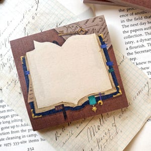 Op de afbeelding: Vierkante bruine en crèmekleurige plaknotities met een open boekontwerp. De notities hebben een donkerblauwe rand met gouden accenten en een kleine ster- en halve maan-bedel. Briefpapier in vintage stijl.