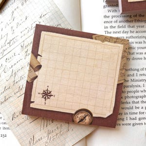 Peut inclure: Un bloc-notes carré de style vintage avec une rose des vents, des lignes quadrillées et des ornements en forme de rouleau. Le bloc-notes est posé sur du papier vieilli avec une écriture manuscrite.