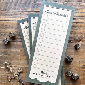 Pode incluir: Três blocos de notas verdes e creme "Roll for Initiative" com um design de lista de verificação, empilhados em uma superfície de madeira. Dados metálicos e uma espada decorativa também estão presentes. Os blocos de notas têm uma borda em forma de castelo.