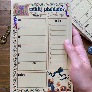 Pu&ograve; includere: Un blocco note per la pianificazione settimanale con un design in stile medievale. La copertina presenta un drago e un castello in fiamme. Il blocco note ha una sezione per ogni giorno della settimana, un elenco di cose da fare e una sezione note.