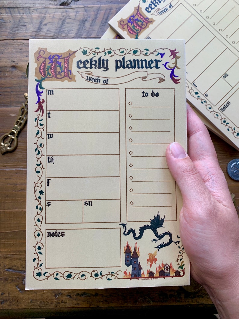 Pu&ograve; includere: Un blocco note per la pianificazione settimanale con un design in stile medievale. La copertina presenta un drago e un castello in fiamme. Il blocco note ha una sezione per ogni giorno della settimana, un elenco di cose da fare e una sezione note.