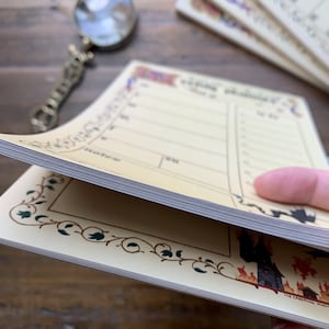 Pu&ograve; includere: Un blocco note giallo con un bordo decorativo e un'illustrazione in bianco e nero di una casa infestata. Il blocco note ha una sezione per "Da fare" e una sezione per "Note".