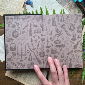 May include: A closed book with a black cover and a beige interior. The interior is covered in a repeating pattern of illustrations of various witchy items, including a cat, a cauldron, a broom, a pumpkin, a key, a mushroom, a candle, a teapot, a pie, a knife, a book, a fern, and a mouse. The book is open to reveal the pattern.