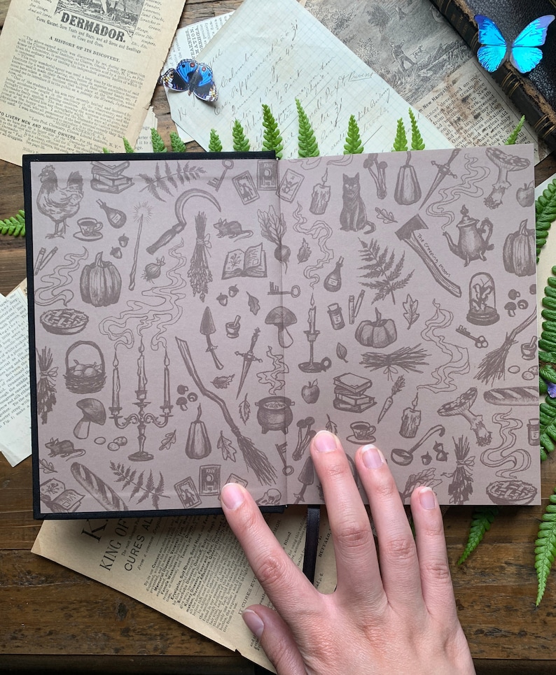 May include: A closed book with a black cover and a beige interior. The interior is covered in a repeating pattern of illustrations of various witchy items, including a cat, a cauldron, a broom, a pumpkin, a key, a mushroom, a candle, a teapot, a pie, a knife, a book, a fern, and a mouse. The book is open to reveal the pattern.
