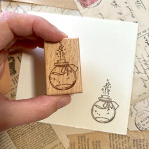 May include: A wooden stamp with a hand-drawn illustration of a glass bottle with a cork stopper and a ribbon. The bottle is filled with a liquid and has stars and sparkles around it. The stamp is perfect for adding a whimsical touch to your crafts and projects.