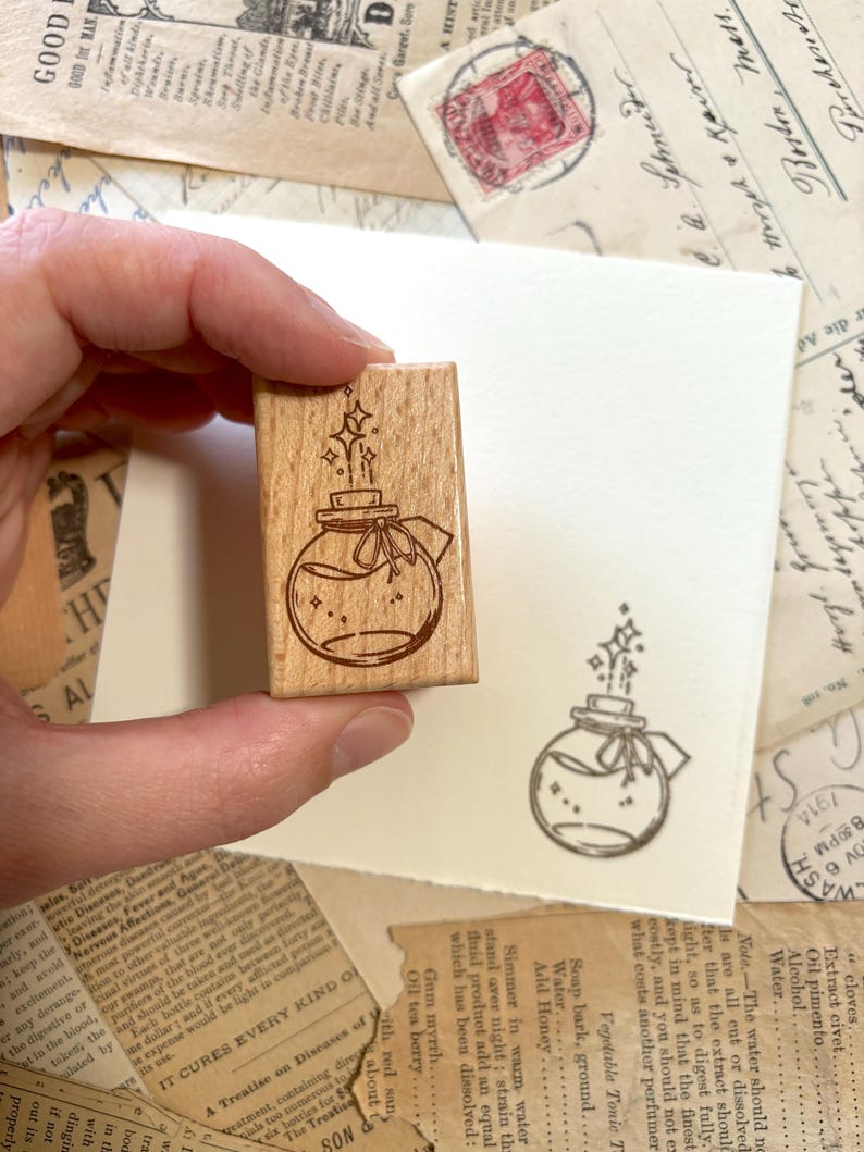 May include: A wooden stamp with a hand-drawn illustration of a glass bottle with a cork stopper and a ribbon. The bottle is filled with a liquid and has stars and sparkles around it. The stamp is perfect for adding a whimsical touch to your crafts and projects.