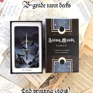 May include: A tarot deck set featuring a black box with the text Anima Mundi Tarot, 78 card deck & guidebook, Megan Wyreweden, and moon graphics. Beside it, an open card shows the Ace of Swords with a sword, crown, and dark mountain landscape.