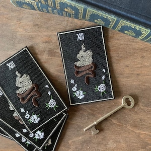 May include: Four black embroidered iron-on patches with white roses and a brown snake with the Roman numeral XIII. The patches are on a wooden surface with a silver key.