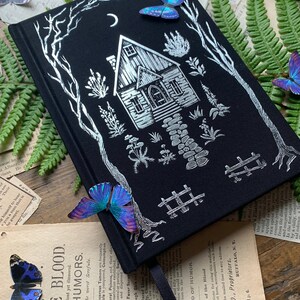 May include: A black journal with a silver foil design of a house in the woods. The house has a crescent moon above it and a path leading up to the front door. There are butterflies on the cover of the journal.