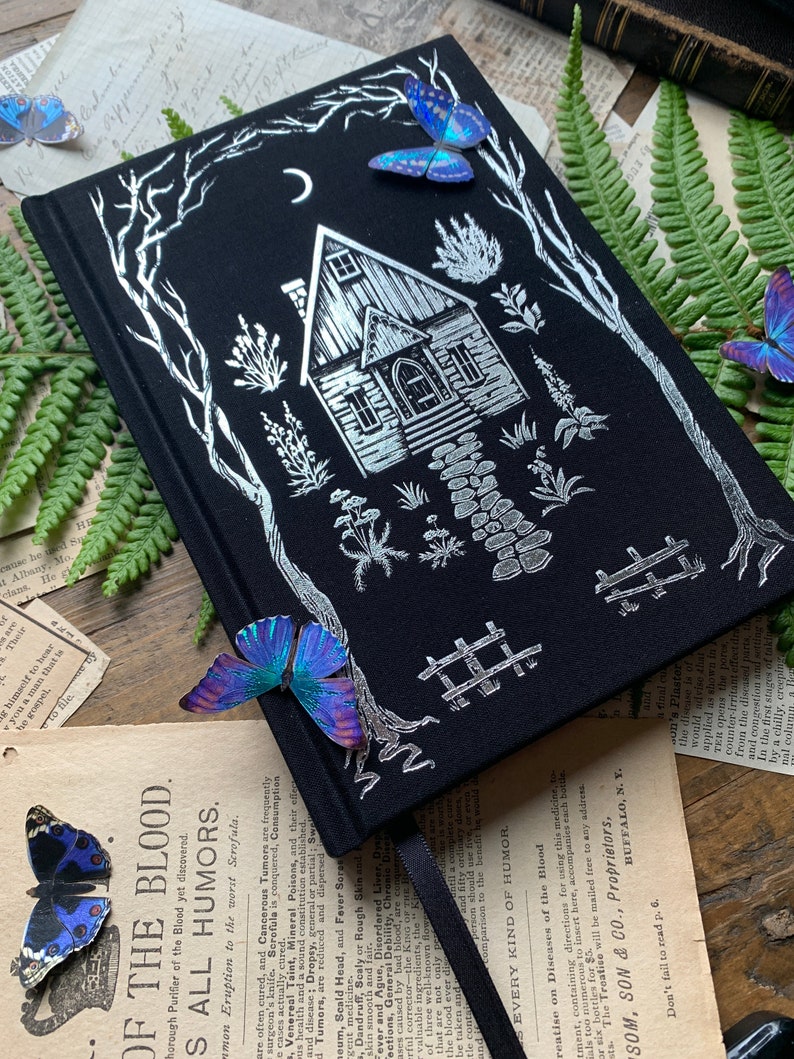 May include: A black journal with a silver foil design of a house in the woods. The house has a crescent moon above it and a path leading up to the front door. There are butterflies on the cover of the journal.