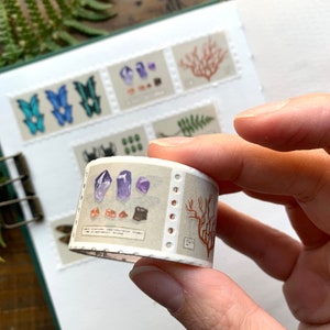 May include: A roll of decorative washi tape featuring illustrations of crystals, coral, and butterflies. The tape is held in a hand, with a notebook and fern in the background. The tape has a light beige background with a perforated edge.