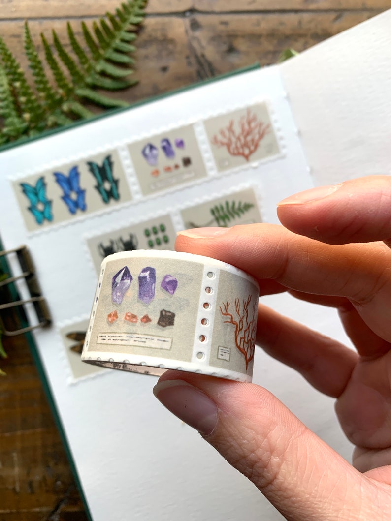 May include: A roll of decorative washi tape featuring illustrations of crystals, coral, and butterflies. The tape is held in a hand, with a notebook and fern in the background. The tape has a light beige background with a perforated edge.