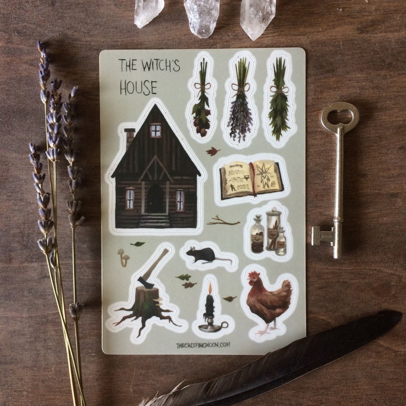 May include: A sticker sheet with a witch's house, a book, a rat, a chicken, an axe, a candle, and a few bottles. The sheet is on a wooden surface with lavender sprigs and crystals.