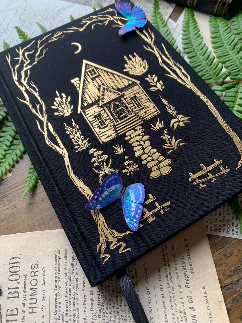 May include: A black hardcover journal with a gold foil design of a house with a crescent moon above it. The house is surrounded by trees and flowers. There are two iridescent blue butterflies on the cover.