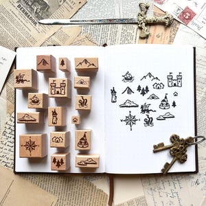 May include: A collection of wooden stamps featuring various icons including a compass, castle, dragon, trees, and clouds. The stamps are arranged on an open notebook page that displays black ink impressions of each design, alongside a metal sword and keys.