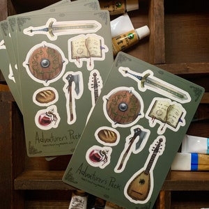 May include: A set of 10 green and white stickers featuring fantasy role-playing game items. The stickers include a sword, shield, axe, lute, book, bread, and a potion. The stickers are labeled "Adventurer's Pack" and "thecreepingmoon.co".