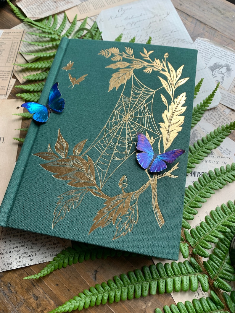 Op de afbeelding: Een donkergroen hardcover dagboek met een gouden folieontwerp van een spinnenweb en bladeren. Twee blauwe vlinders en een paarse vlinder zijn op de omslag bevestigd.