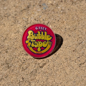 May include: A round, red plastic container of Bubble Tape bubblegum. The lid features yellow text that reads 6 FEET OF Bubble Tape and ORIGINAL BUBBLEGUM in smaller red letters. The container has a clear plastic rim and a flat, circular top surface.