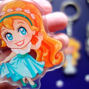 May include: A clear acrylic keychain featuring a cartoon girl with orange hair, blue eyes, and a turquoise dress. The girl is waving and smiling. Other keychains are visible in the background.