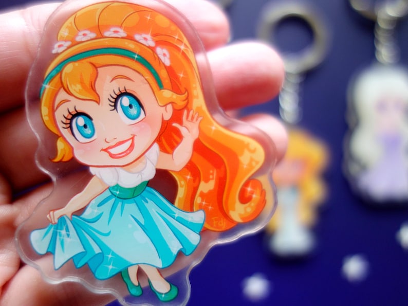May include: A clear acrylic keychain featuring a cartoon girl with orange hair, blue eyes, and a turquoise dress. The girl is waving and smiling. Other keychains are visible in the background.