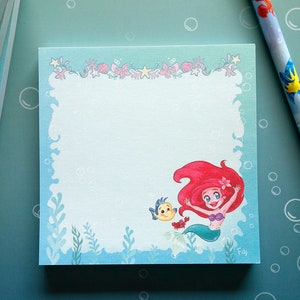 Peut inclure: Un bloc-notes bleu avec une image de dessin animé d'Ariel de La Petite Sirène. Ariel nage avec Polochon et un crabe. Le bloc-notes a un fond blanc et une bordure bleue avec un motif sur le thème de la mer.